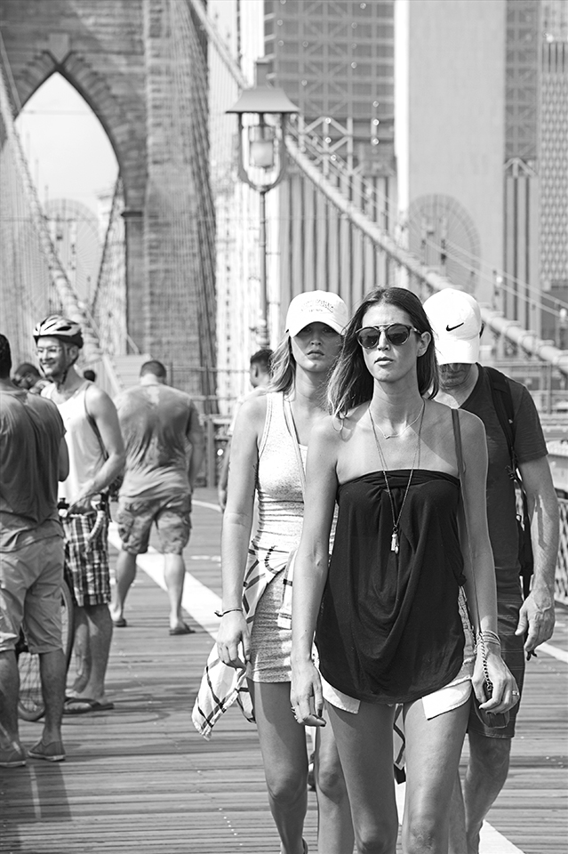 Black and white image of pedestrians walking on the upper-level of the Brooklyn Bridge, it’s summer time, everyone in this image is wearing shorts