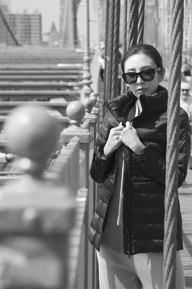 Black and white image of a woman wearing dark sunglasses, she has her hands on the lapels of her jacket and she is facing the viewer over her left shoulder on the upper-level of the Brooklyn Bridge