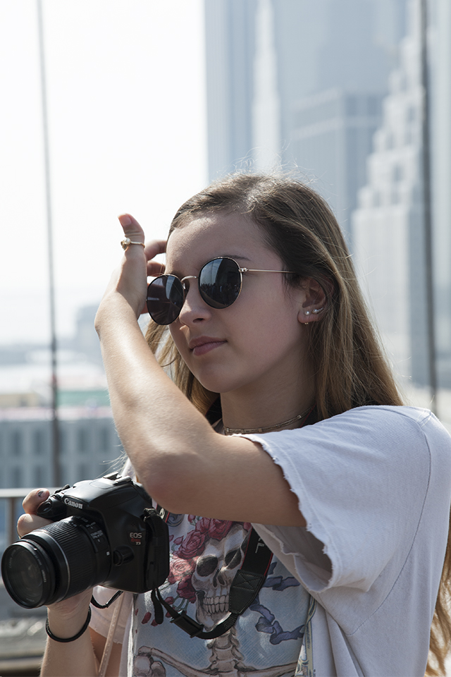 Young woman wearing a Grateful Dead t-shirt, dark, round sunglasses and holding a Cannon camera, upper-level of the Brooklyn Bridge