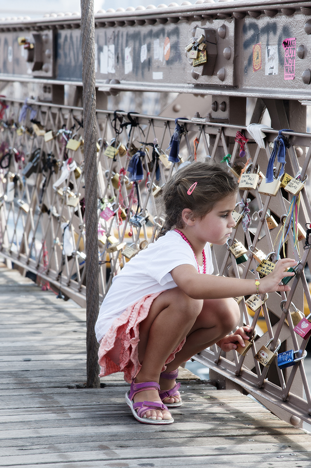 A young girl with brown hair in pig-tails wearing a pink shirt and white top is taking a closer look at a love lock attached to a fence on the Brooklyn Bridge