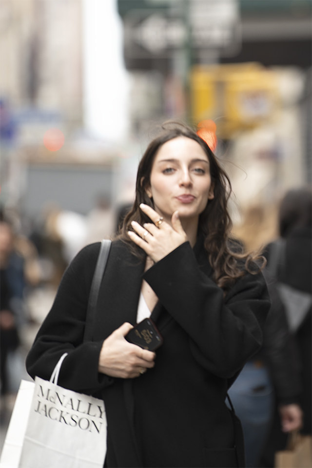 Woman with long brown shoulder length hair, wearing a black coat, an holding a cell phone in her righthand, with a McNally Jackson shopping bag on her right arm as she looks at the viewer, East Village, Manhattan