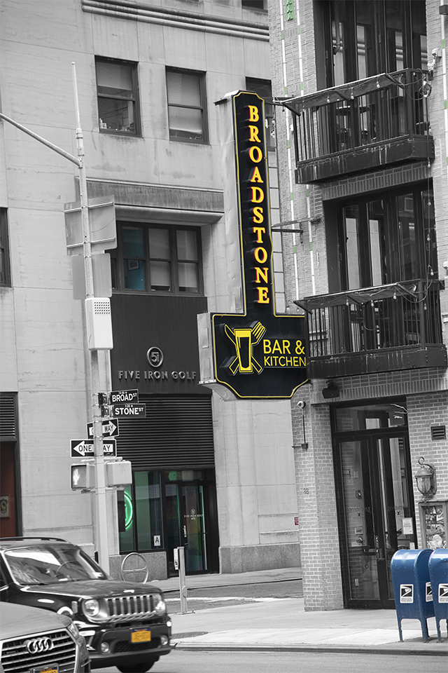 Black and white image, lower Manhattan, with selective parts the image colorized, i.e. license plates, a storefront illuminated by green neon sign, the store sigh “Broadstone Bar and Kitchen" sign, and 2 mailboxes on the corner