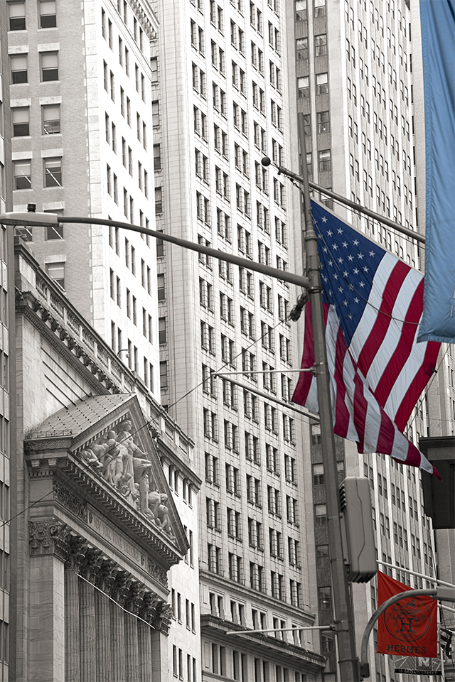 Black and white image with colorized parts. The image is of the New York Stock Exchange Broad Street Façade, the exchange is on the viewers lefthand side of the image. On the viewers righthand side of the image there are to flags that have been colorized